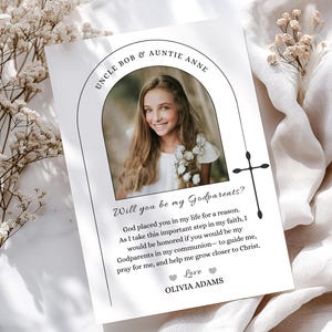 May include: A white card with a photo of a girl, text that reads "Uncle Bob & Auntie Anne" and "Will you be my Godparents?" The card includes a cross graphic and the name "Olivia Adams". The card is surrounded by white flowers.