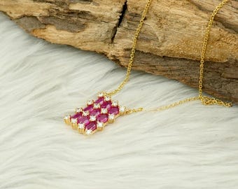 Op de afbeelding: Een goudkleurige ketting met een hanger met rode en witte edelstenen. De hanger heeft een geometrisch ontwerp. De ketting wordt getoond op een witte, zachte ondergrond met een houten achtergrond.