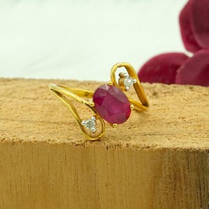 May include: A gold ring featuring a large, oval-cut ruby gemstone as the centerpiece. The ring has a delicate design with two smaller diamonds and a unique, swirling band. The ruby is a deep, rich red color.