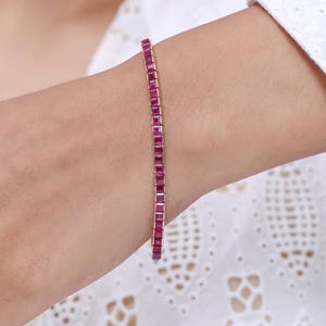 Pode incluir: Uma pulseira delicada com uma série de pedras preciosas vermelhas vibrantes, de corte quadrado. As pedras são colocadas juntas, criando uma linha contínua de cor. A pulseira é uma joia clássica.