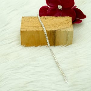May include: A silver tennis bracelet featuring multiple round, clear gemstones. The bracelet is presented on a wooden block with a red flower in the background, against a white, fluffy surface. It fastens with a clasp.