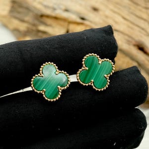 May include: A pair of clover-shaped earrings. Each earring features a vibrant green malachite stone set in a gold-colored beaded frame. The earrings are displayed on a black surface, showcasing their design and color.