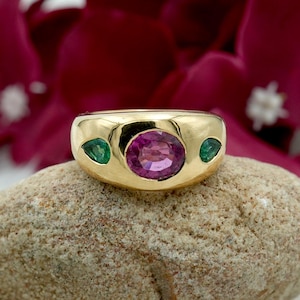 May include: A gold ring featuring a large oval-cut pink gemstone in the center, flanked by two smaller, triangular green gemstones. The ring is set on a light brown stone, with a blurred red floral background.