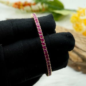 Puede incluir: Una pulsera dorada con una fila de gemas rojas vibrantes. La pulsera se muestra sobre un fondo negro, resaltando el color de las gemas y el engaste dorado. Las gemas están espaciadas uniformemente, creando una línea continua.