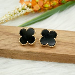 May include: A pair of black clover-shaped stud earrings with a gold beaded border. The earrings are displayed on a wooden surface, with blurred flowers in the background. The earrings have a classic and elegant design.