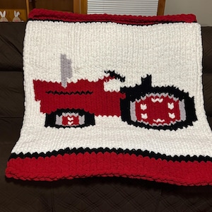 May include: A chunky knit blanket featuring a white background with a large red and black tractor graphic in the center. The blanket has a thick red border at the top and bottom, accented by thin black stripes, and is made of soft, thick yarn.