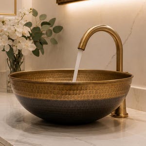 May include: A hammered brass sink with a dark blue band sits on a marble countertop. A gold faucet pours water into the sink. A vase of white flowers and a gold-framed mirror are in the background.