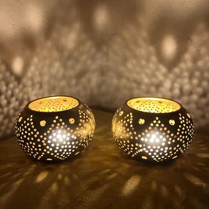 May include: Two decorative, dark brown candle holders with intricate dot patterns. The candle holders are round with a series of small holes that allow light to shine through. The warm light creates a cozy ambiance.