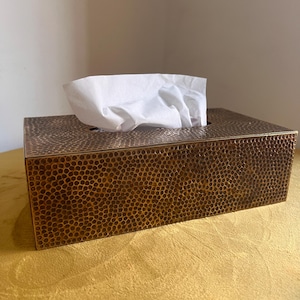 Może przedstawiać: Prostokątne pudełko na chusteczki o teksturowanej, brązowej powierzchni. Pudełko ma wzór z małych, równomiernie rozmieszczonych kół. Białe chusteczki są wyciągnięte z górnego otworu. Pudełko stoi na złotej powierzchni.
