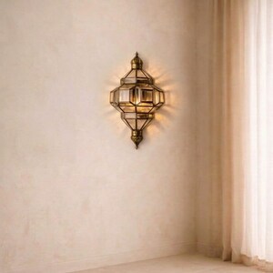 May include: An ornate, antique-style wall sconce with a geometric design. The sconce is made of brass-colored metal and features multiple glass panels. It is mounted on a beige wall, with a soft light emanating from within. The overall aesthetic is vintage and elegant.