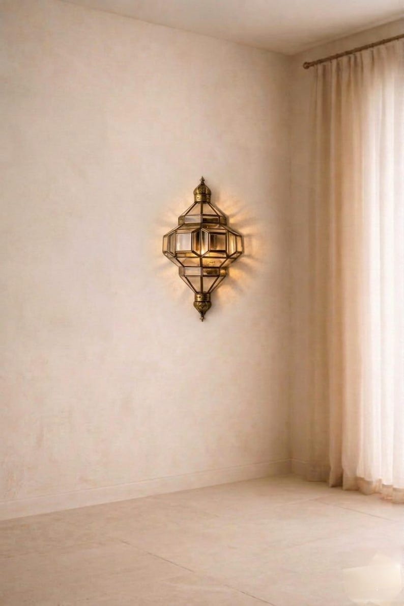 May include: An ornate, antique-style wall sconce with a geometric design. The sconce is made of brass-colored metal and features multiple glass panels. It is mounted on a beige wall, with a soft light emanating from within. The overall aesthetic is vintage and elegant.