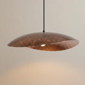 May include: A contemporary pendant light in a copper finish. The light features a broad, curved shade with a textured surface and a single bulb. It is suspended from a black cord against a neutral backdrop.