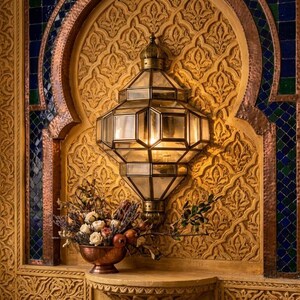 May include: An ornate wall sconce with a geometric glass shade is mounted on a carved, golden wall. A copper bowl with flowers and fruit sits below the light fixture. The wall features intricate patterns and mosaic accents.