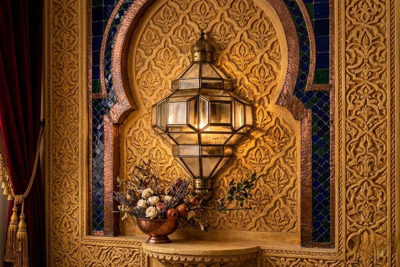 May include: An ornate wall sconce with a geometric glass shade is mounted on a carved, golden wall. A copper bowl with flowers and fruit sits below the light fixture. The wall features intricate patterns and mosaic accents.