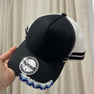May include: Black and white trucker cap with a beaded trim. The cap has a black front panel, a white mesh back, and a curved peak. The peak is decorated with blue and white beads. The cap has the text "AUSSIE RETRO TRUCKER" on the front.