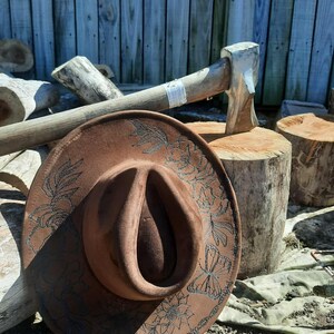 May include: A brown cowboy hat with floral and butterfly patterns. The hat is placed on a wooden surface, with an axe and logs visible. The hat's brim features detailed decorative designs.