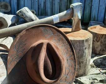 May include: A brown cowboy hat with floral and butterfly designs. The hat is resting on a wooden surface with an axe and logs in the background. The hat's brim is decorated with intricate patterns.
