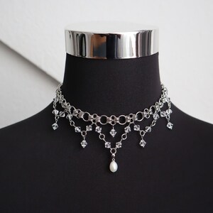 May include: Silver-toned choker necklace with multiple strands and dangling crystal accents. A single pearl pendant hangs from the center. The necklace is displayed on a black mannequin.