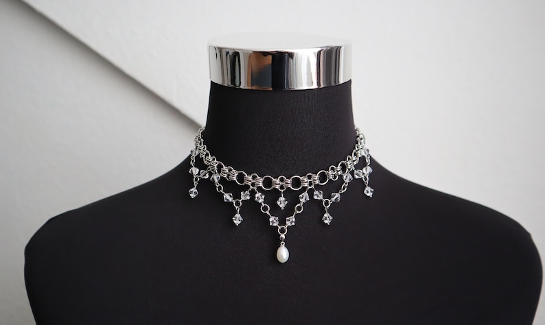 May include: Silver-toned choker necklace with multiple strands and dangling crystal accents. A single pearl pendant hangs from the center. The necklace is displayed on a black mannequin.