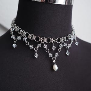 May include: Silver-toned choker necklace with multiple linked chains and dangling clear crystals. A single white pearl pendant hangs from the center. The necklace is displayed on a black mannequin.