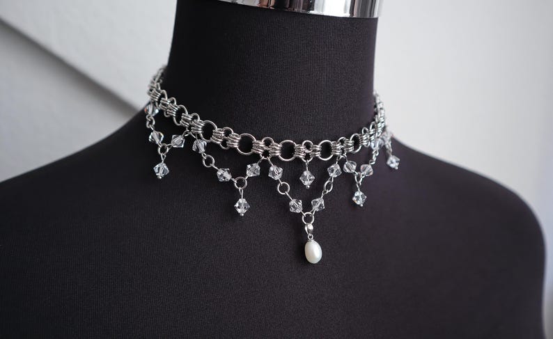 May include: Silver-toned choker necklace with multiple linked chains and dangling clear crystals. A single white pearl pendant hangs from the center. The necklace is displayed on a black mannequin.