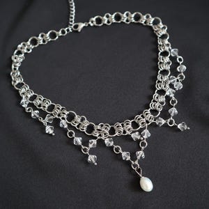 May include: A silver-toned necklace with a chainmail design. Clear crystal beads and a single white pearl dangle from the necklace. The necklace is displayed against a black background.