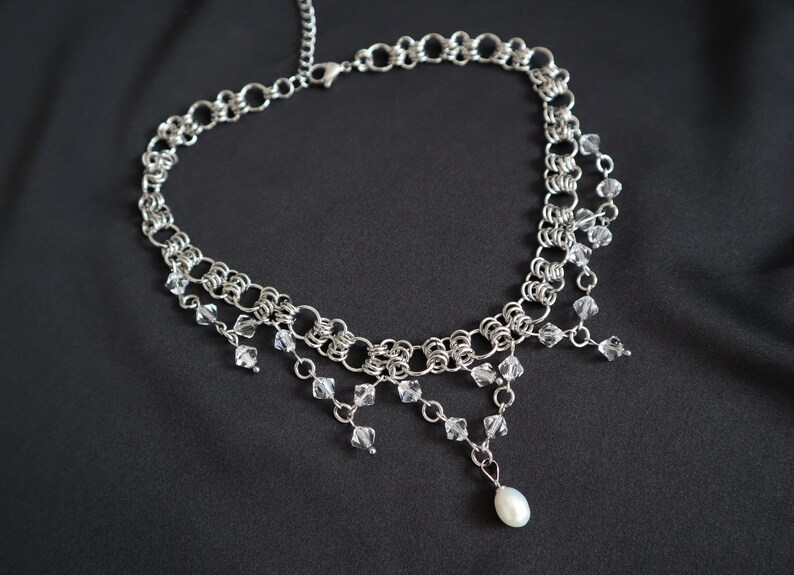 May include: A silver-toned necklace with a chainmail design. Clear crystal beads and a single white pearl dangle from the necklace. The necklace is displayed against a black background.