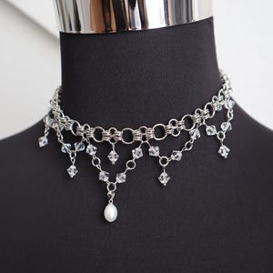 May include: A silver-toned choker necklace with multiple layers of chain and dangling clear crystals. A single white pearl hangs from the center. The necklace is displayed on a black mannequin.