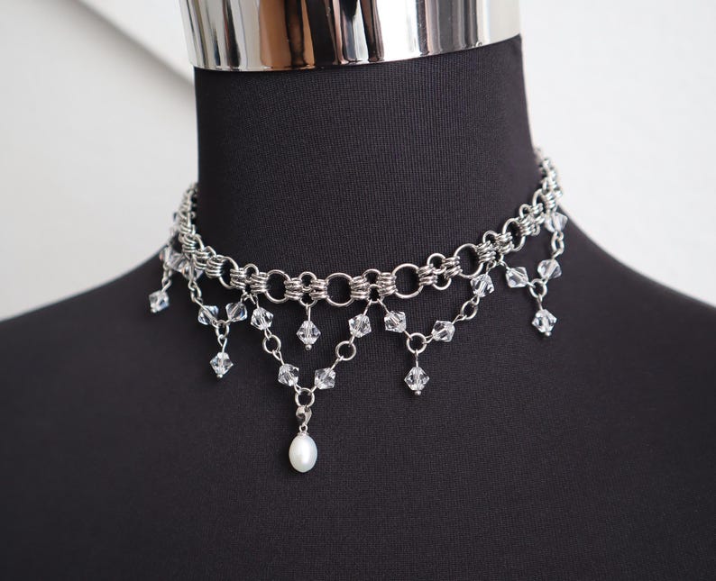 May include: A silver-toned choker necklace with multiple layers of chain and dangling clear crystals. A single white pearl hangs from the center. The necklace is displayed on a black mannequin.