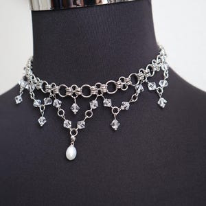 May include: A silver-toned choker necklace with a layered design. The necklace features multiple strands of linked metal rings and dangling clear crystals. A single white pearl pendant hangs from the center, adding a touch of elegance.
