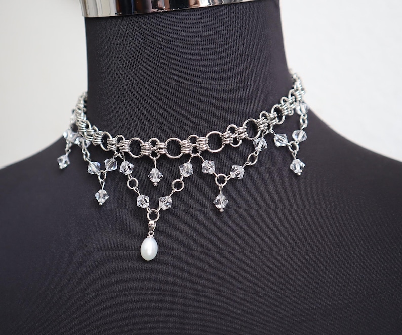 May include: A silver-toned choker necklace with a layered design. The necklace features multiple strands of linked metal rings and dangling clear crystals. A single white pearl pendant hangs from the center, adding a touch of elegance.
