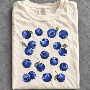 May include: A cream-colored short-sleeve t-shirt featuring a repeating pattern of dark blue blueberries with small green leaves. The shirt has a crew neckline and a Comfort Colors brand label printed on the inside of the collar with size L G G.