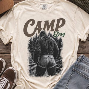 May include: A cream-colored t-shirt featuring a graphic of a hairy creature standing in a forest, wearing denim shorts. The text "CAMP Day" is printed above the creature in brown and green. The shirt is surrounded by a brown cap, a green beanie, brown sneakers, and blue jeans.