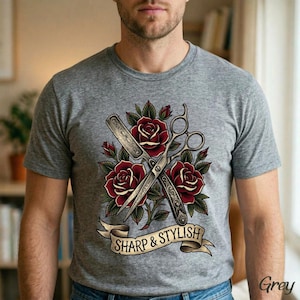 May include: Gray t-shirt featuring a barber-themed design. The graphic includes crossed barber scissors and a straight razor, surrounded by red roses and green leaves. A banner below reads "Sharp & Stylish".