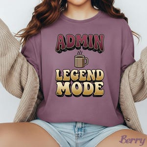 May include: Berry-colored t-shirt with the text "ADMIN LEGEND MODE" in a retro font. A coffee cup graphic is between the words. The shirt is a solid color and has a relaxed fit.