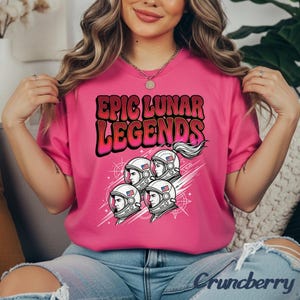 Puede incluir: Una camiseta rosa brillante con el texto "EPIC LUNAR LEGENDS" en un estilo retro. El diseño incluye cuatro ilustraciones de astronautas con detalles de la bandera estadounidense en sus cascos. La camiseta es de un color vibrante.
