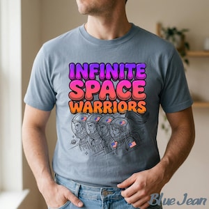 Puede incluir: Una camiseta azul claro con el texto "INFINITE SPACE WARRIORS" en una fuente colorida y de burbujas. Debajo, un dibujo lineal de cuatro astronautas con detalles de la bandera estadounidense en sus cascos.