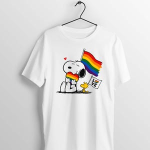 Può includere: Una t-shirt bianca a maniche corte con una grafica di Snoopy che tiene in mano un cuore arcobaleno, una grande bandiera arcobaleno e Woodstock accanto a una piccola bandiera bianca con il testo LOVE e un'icona a cuore arcobaleno.