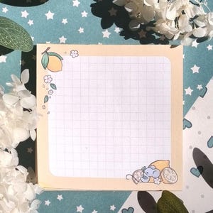 Może przedstawiać: Kwadratowy notatnik z jasnożółtą obwódką i wzorem kratki. Notatnik jest ozdobiony cytrynowym i kwiatowym wzorem w rogach. Tło jest jasnoniebieskie z białymi gwiazdami i kwiatowymi akcentami.