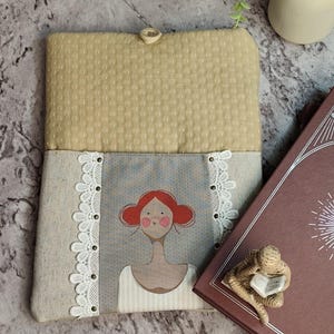 Puede incluir: Una funda de libro beige acolchada con cierre de botón. La funda presenta un panel gris con una ilustración caprichosa de una mujer con cabello rojo y un vestido blanco, enmarcada por un ribete de encaje blanco. Se ve un libro marrón.