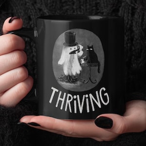May include: Black ceramic mug featuring a whimsical illustration of a ghost in a top hat and a black cat. The word "THRIVING" is printed in white below the illustration. The mug is held by a person wearing a black sweater.
