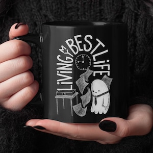 May include: Black ceramic mug with the words "LIVING BEST LIFE" in white, along with a clock and a cartoon ghost. The mug is held by a person wearing a black sweater.