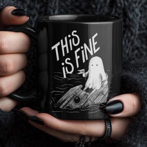 May include: Black ceramic mug with the phrase "THIS IS FINE" in white lettering. The mug features a cartoon ghost sitting on a sinking ship. The mug has a black handle and is held by a person wearing a black sweater.