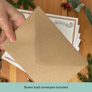 May include: A stack of white cards with red and green border designs, topped with a brown kraft envelope. The text "the revels are upon us" is visible on the cards. The image is set on a wooden surface with festive greenery.