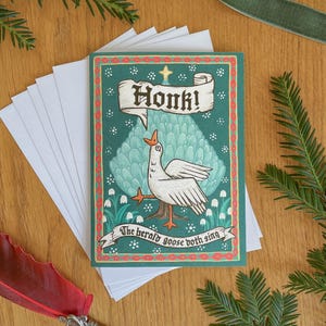 以下が含まれることがあります： 「Honk!」の文字が入ったバナーを持つガチョウのイラストが描かれた、気まぐれなグリーティングカード。カードは、赤い縁取りのあるティール色の背景で、「The herald goose both sing」というテキストが入っています。白い封筒がカードの隣に積み重ねられています。
