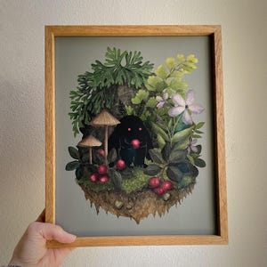 May include: A framed art print featuring a black creature with glowing red eyes holding a red berry. The creature is surrounded by green ferns, light purple flowers, brown mushrooms, and red berries on a patch of earth against a gray background.