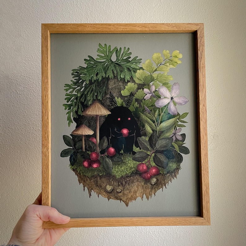 May include: A framed art print featuring a black creature with glowing red eyes holding a red berry. The creature is surrounded by green ferns, light purple flowers, brown mushrooms, and red berries on a patch of earth against a gray background.
