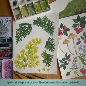 May include: Two sheets of watercolor paper featuring hand-painted botanical illustrations of green ferns, yellow foliage, brown mushrooms, red berries, and purple flowers, accompanied by watercolor paint palettes and brushes on a wooden surface.