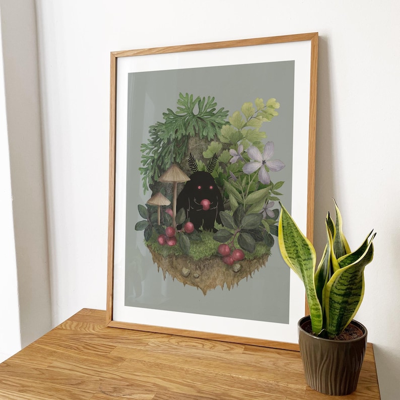 May include: A framed art print featuring a dark, fuzzy creature with glowing pink eyes holding a red berry, surrounded by green foliage, mushrooms, and purple flowers on a floating patch of earth against a solid light gray background.