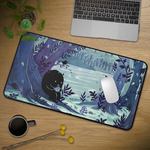 May include: A rectangular mouse mat with a forest illustration in blue and purple hues. A black creature is shown holding a cup, with a river and trees in the background. A white mouse is placed on the mat.